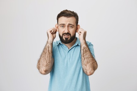 Adult bearded man with gloomy smile stretching ears, imitating sad monkey, over gray background. Guy...