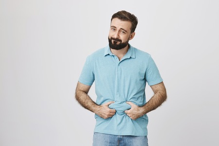Picture of a bearded handsome man showing his beer belly over white background. Person is bothered...