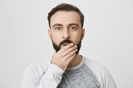Picture of handsome man with a beard expressing surprise covering his mouth with hand over white background. Coworker just told an interesting gossip about their employer.の写真素材