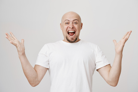 Studio portrait of overwhelmed and excited bald caucasian man with beard shouting of happiness and spreading hands, showing his joy because of success over gray background. Guy became fatherの写真素材
