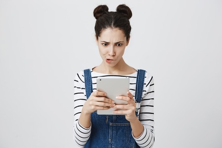 Portrait of cute european girl with trendy two buns hairstyle, wearing overalls and looking excited or interested at tablet screen, standing over gray background. Woman looks who liked her boyfriendの写真素材