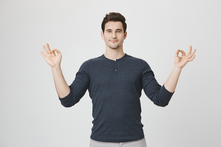 Calm and concentrated young handsome model, standing with raised hands and zen gesture, acting like meditating or doing yoga while standing over gray background. Guy repeats after his trainerの写真素材