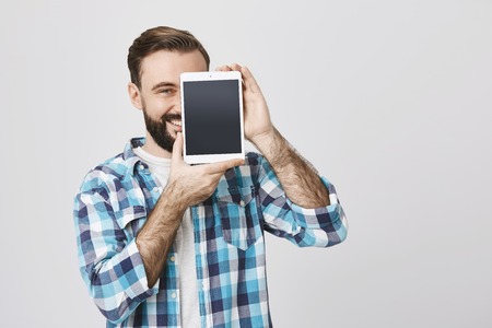 Portrait of male bearded shop assistant covering half-face with tablet to advertise it while smiling...