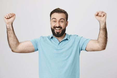 Happy bearded man raising his hands showing victory gesture near white wall. Handsome guy just won in a table game he played with friends and now is on cloud nine, looking happy like a child.の写真素材