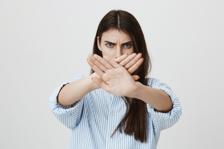 Waist-up portrait of anxious and serious brunette crossing hands in stop or enough gesture, looking angry at camera while standing over gray background. Girl orders to stop harassing herの写真素材