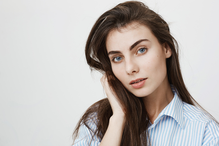 Close-up portrait of charming sensual girlfriend looking dreamily at camera while touching hair and standing over gray background. Woman sits in restaurant and looks through windowの写真素材