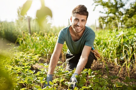 Healthy lifestyle. Countryside life. Close up outdoors portrait of young attractive bearded caucasian farmer smiling, spending morning in garden near house, picking cropの写真素材