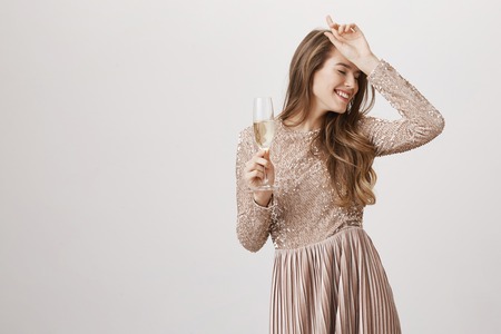 Indoor shot of positive attractive young female in evening dress with glass of champagne, holding hand on forehead and smiling, standing with closed eyes over gray background. Girl dances sensuallyの写真素材