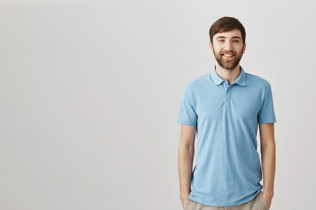 Friendly neighbor asks how was your day. Portrait of good-looking slim man with beard standing in blue polo shirt over gray background with hands in pockets, smiling broadly at cameraの写真素材