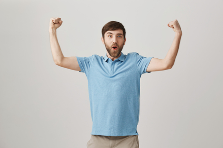 Emotive young caucasian man with beard and moustache lifting hands and showing muscles, expressing excitement and positive emotions while standing over gray background. Guy happy he got promotedの写真素材