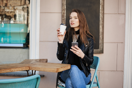 Girl enjoying warm and refreshing cup of coffee during lunch. Busy and successful woman sitting in cafe, drinking tea and dreaming about warm weather, holding smartphone to answer important callの写真素材