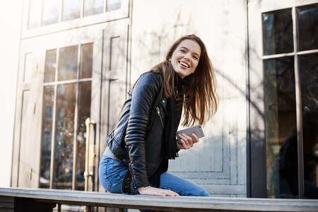 City with positive citizens. Beatiful trendy woman in leather jacket sitting near cafe, leaning on bench, looking aside, holding smartphone, waiting for friend. Urban girl enjoys being outsideの写真素材