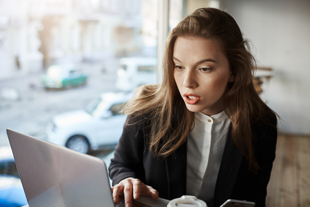 Who left bad reply about my company. Indoor portrait of bothered and confused stylish girl sitting in cafe, working with laptop, looking at screen with surprised expressionの写真素材