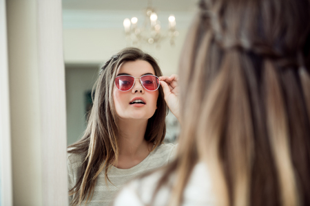 Close-up portrait of attractive modern european woman in optician store standing near mirror and trying on stylish sunglasses, deciding between few pairs and consulting with sales assistantの写真素材