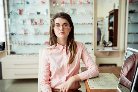 Glasses is not just for sight it is for style. Serious and confident caucasian woman in formal outfit sitting in optician store in presctibed glasses, looking at camera while waiting for check-upの写真素材