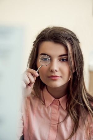 Horizontal portrait of good-looking focused woman on appointment with ophthalmologist holding lense and looking through it while trying to read word chart to check vision. Eyecare and health conceptの写真素材