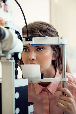 Horizontal portrait of focused european woman testing sight while looking through microbioscope, sitting in specialist office, wanting to pick appropriate glasses to see betterの写真素材