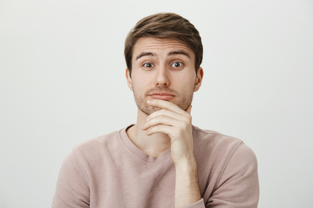 We should reconsider it together. Portrait of handsome troubled european man lifting eyebrow and holding hand on chin, puckering while thinking, standing over gray background cluelessの写真素材