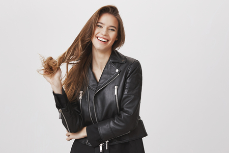 Beautiful and rebel woman playing with hair. Portrait of charming modern european girl in leather jacket touching strands and smiling broadly, having chat with handsome barista while waiting coffeeの写真素材