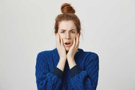Oh dear, it is disaster. Worried beautiful modern girl with red hair and freckles in bun and sweater holding hands on cheeks and looking with anxious expression at camera, seeing terrifying sceneの写真素材