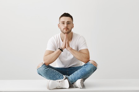 Time for some procrastination. Portrait of attractive dreamy guy with stylish haircut and glasses sitting on floor with crossed legs, leaning chin on clasped hands, being over gray backgroundの写真素材