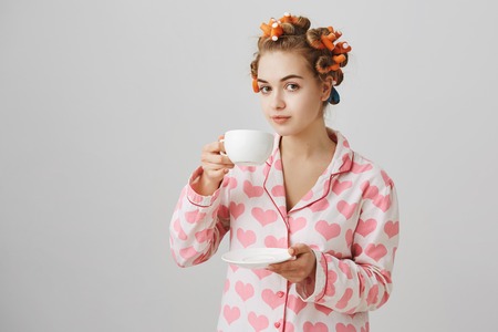 Housewife making procedures on weekend. Portrait of cute and funny european woman in hair curlers and nightwear with heart pattern, holding cup of coffee or tea, drinking while standing over gray wallの写真素材