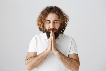 God has answers. Portrait of calm and focused eastern man with curly hair and beard holding hands in pray near mouth, closing eyes while begging or hoping for better, standing over gray wallの写真素材