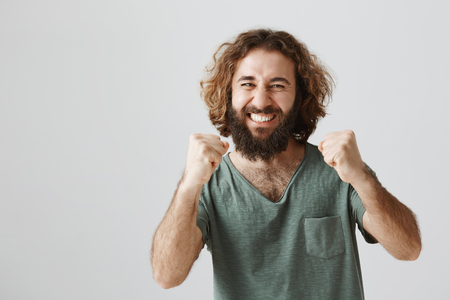 I was sure in victory. Portrait of happy celebrating easter man with curly hair and beard raising fists and shaking them in success gesture, smiling broadly and being proud of friend who achieved goalの写真素材