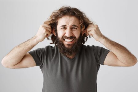 Ears are tired from hearing lies. Studio shot of bothered and depressed mature guy with beard and curly hair pulling ears and grimacing from pain, being unhappy and stressed over gray backgroundの写真素材