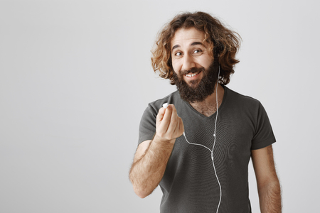 Check out great sound of this earphones. Pleased attractive curly-haired eastern man pulling headphone at camera with broad smile, suggesting coworker to listen new song, standing over gray backgroundの写真素材