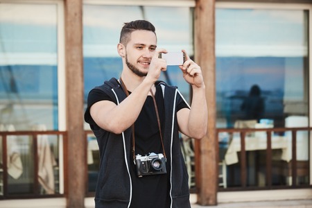 Photographers love this smartphone. Outdoor portrait of handsome european photographer in trendy clothes taking photo on phone while wearing camera on neck, smiling, having flair for photographyの写真素材