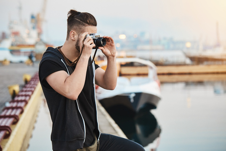 Capturing beautiful sunset. Dreamy creative european photographer in stylish outfit standing in harbour, taking picture of sea or yacht, gathering shots for his studio, being focused on shotsの写真素材