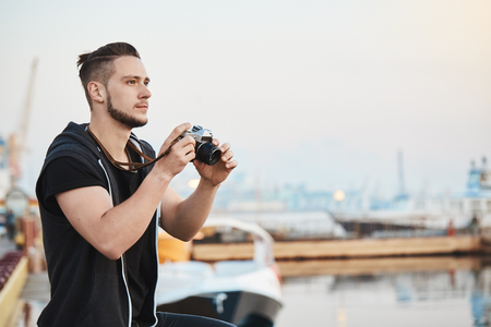Dreamy talented cameraman impressed with beauty of nature while taking photos on camera, looking at blue sky, standing in harbour near sea. Handsome guy capturing nice shots of seaview while walkingの写真素材