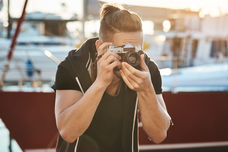 Cameraman trying hold still not to scare birds. Portrait of focused young male photographer looking through camera and frowning, being focused on model during photo session near seashore in harbourの写真素材