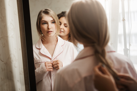 Close-up portrait of two beautiful women at home. Attractive young blonde standing near mirror, changing clothes from pyjamas and waiting while mother combing braid. Typical cozy morning in familyの写真素材