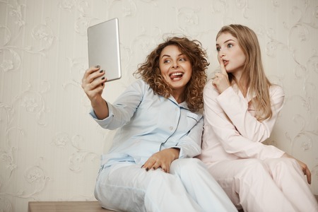 Daughter and mother making family photo while relaxing at home. Emotive beautiful mom taking selfie with cute bestie while both in nightwear, making faces and shh gesture as if having secretの写真素材