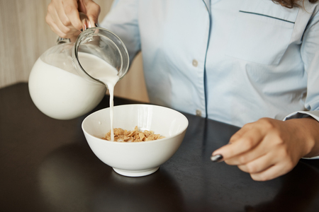 Girlfriend preparing simple breakfast in morning. Cropped shot of woman in nightwear pouring milk in bowl with cereals, wanting to eat fast and getting dressed to go to officeの写真素材