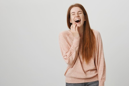 Charming sincere redhead european student with freckles laughing out loud with closed eyes while touching face with hand and binding towards camera while standing over gray background.の写真素材