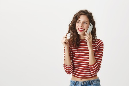 Keep receiving positive feedback from my clients. Portrait of happy satisfied stylish woman talking on smartphone while touching strand of hair, being focused on conversation, smiling over gray wallの写真素材