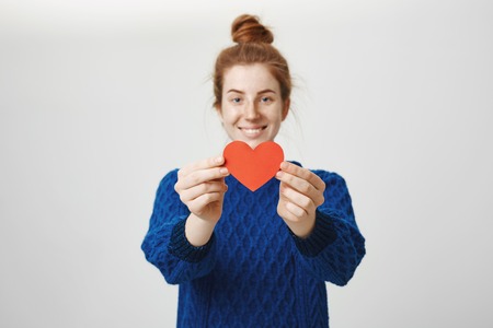 Keep it safe in your hands. Portrait of charming young woman in love pulling paper heart towards camera and smiling cheerfully while offering to start relationship to person she admiresの写真素材