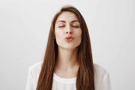 Give your girlfriend tender mwah. Indoor shot of cute romantic european female closing eyes and pulling head towards camera with puckered lips, waiting for kiss with smile, standing over gray wallの写真素材