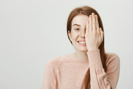 Smiling playful caucasian redhead with cute freckles hiding half of face behind palm, smiling cheerfully and standing against gray background.の写真素材