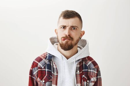 Studio shot of good-looking pierced urban male skater with stylish haircut and beard showing tongue,...