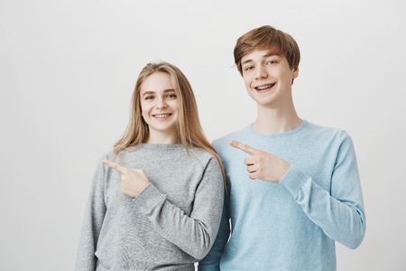 Attractive brother and sister with fair hair, pointing left with index fingers and smiling broadly, indicating at cafe over gray backgroundの写真素材