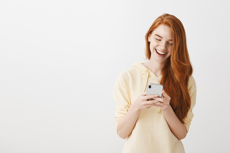 Pleased attractive fashion blogger with natural red hair, holding smartphone, typing message and laughing from happiness, being in good mood over gray wallの写真素材