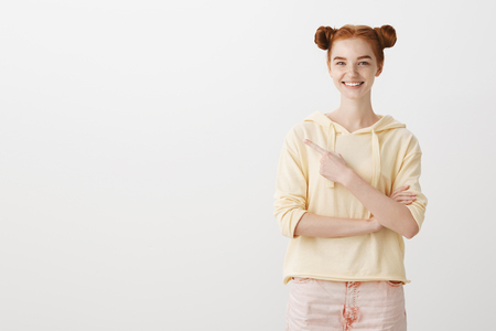 Portrait of charming young caucasian model with ginger hair grinning, expressing positive attitude while pointing left with forefinger, smiling over gray wallの写真素材
