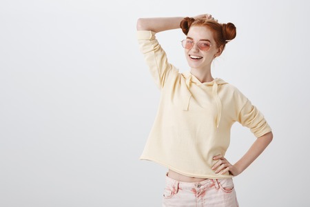 Playful redhead woman with buns hairstyle in pink trendy eyewear posing with hands on hip and head, smiling broadly, feeling confidentの写真素材