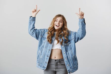 Positive female blogger in fashionable denim jacket and cropped top, raising index fingers and pointing up with pleased excited expressionの写真素材