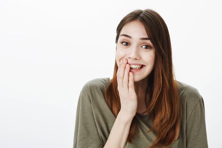 Portrait of happy grinning cute woman in dark-green t-shirt, smiling broadly, holding palm on mouth,...