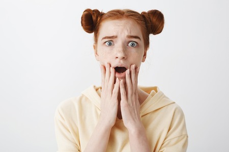 Indoor shot of anxious upset girl with red hair and buns hairstyle dropping jaw, holding fingers near mouth, frowning, being nervous and sympathizingの写真素材